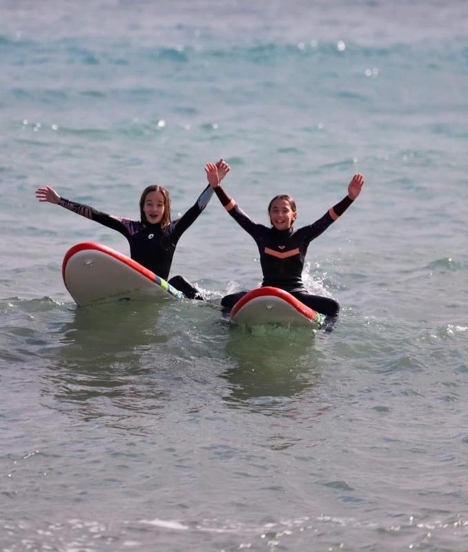 Dos chicas levantando los brazos sentadas en sus tablas de surf en el mar en el campamento verano alicante