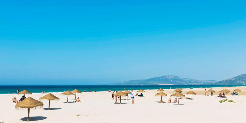 Playa azul, montañas y sombrillas sobre arena blanca en el campamento de verano en Cádiz