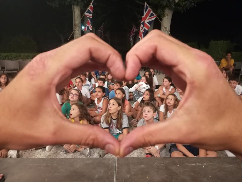 Grupo de niños atendiendo a las funciones de actuacion mientra otro les enmarca con las manos formando un corazon en el campamento de verano en tarragona