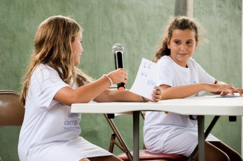 dos niñas sentadas con un micrófono presentando un concurso del campamento baile