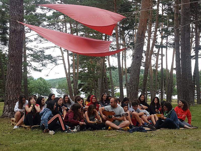Grupo de jóvenes sentados en el césped escuchando a uno de los participantes del campamento de verano en Soria tocando la guitarra