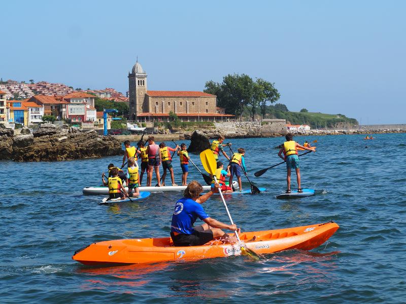 Jóvenes remando sus tablas de SUP mientras el monitor les vigila desde el kayak en el campamento de verano en Asturias
