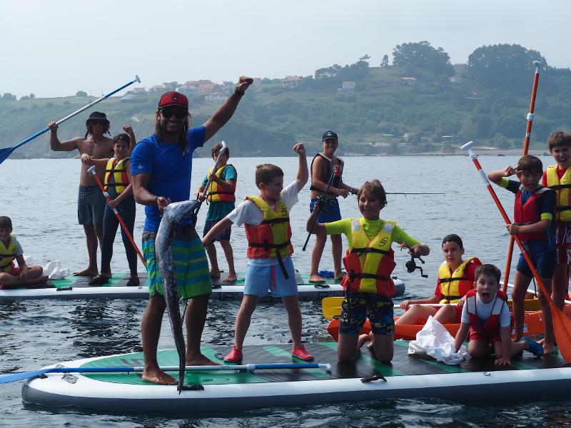 Grupo de niños celebrando porque un monitor ha pescado a un pez grande durante las clases de SUP del campamento náutico en Asturias