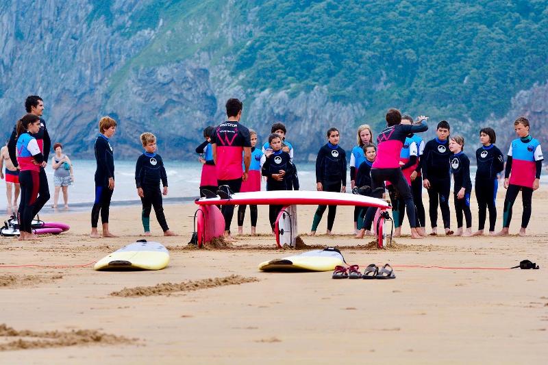 Grupo de niños haciendo un circulo alrededor de los monitores del campamento que explican la clase de surf del día en la playa