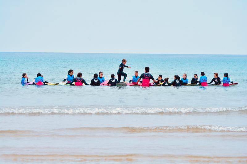 Grupo de jóvenes haciendo una línea en el mar con sus tablas de surf mientras uno a uno tienen que ir caminando encima de las tablas en el campamento