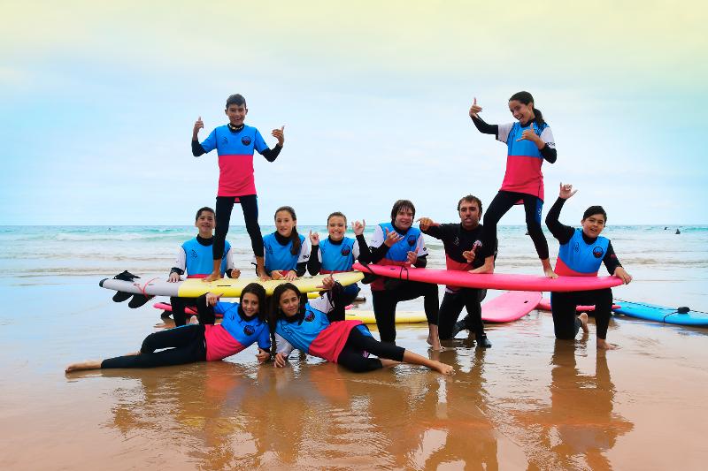 Dos niñas subidas a las tablas de surf mientras el resto de jóvenes las sujetan en la orilla de la playa en el campamento en Cantabria