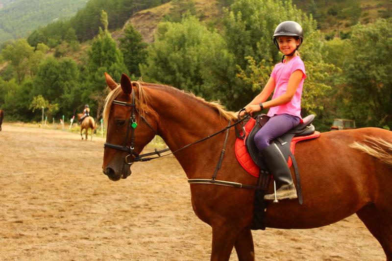 una niña montando a caballo sonriendo a camara en un campamento hipico
