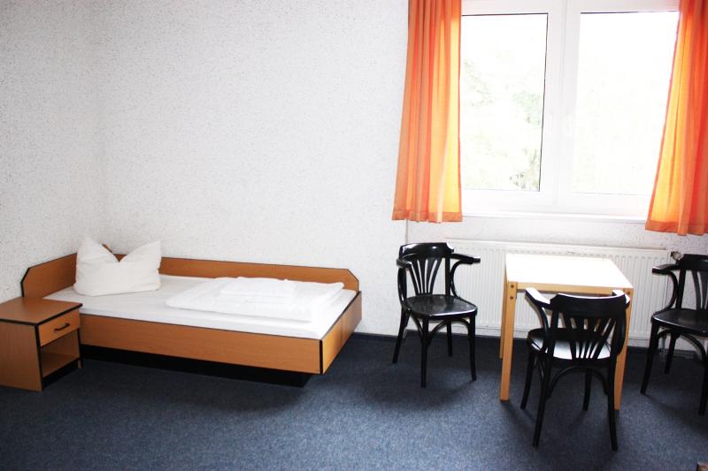 Habitación individual con cama, escritorio, sillas, cómoda y ventana en el campamento de verano en alemania