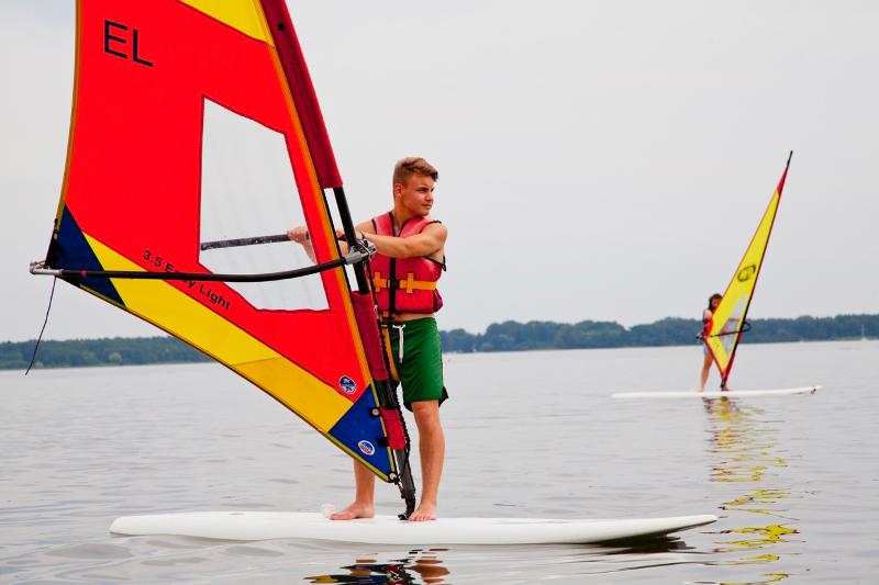 Chico sobre una tabla de windsurf sobre el agua en un lago en campamento de verano en aleman