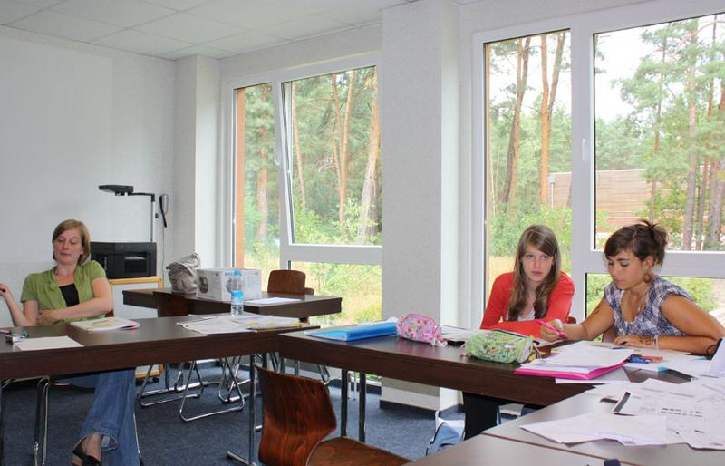 aula de trabajo con dos alumnas y una profesora con papeles y material de estudio en la mesa, detrás hay ventanas grandes hacia la naturaleza en campamento aleman