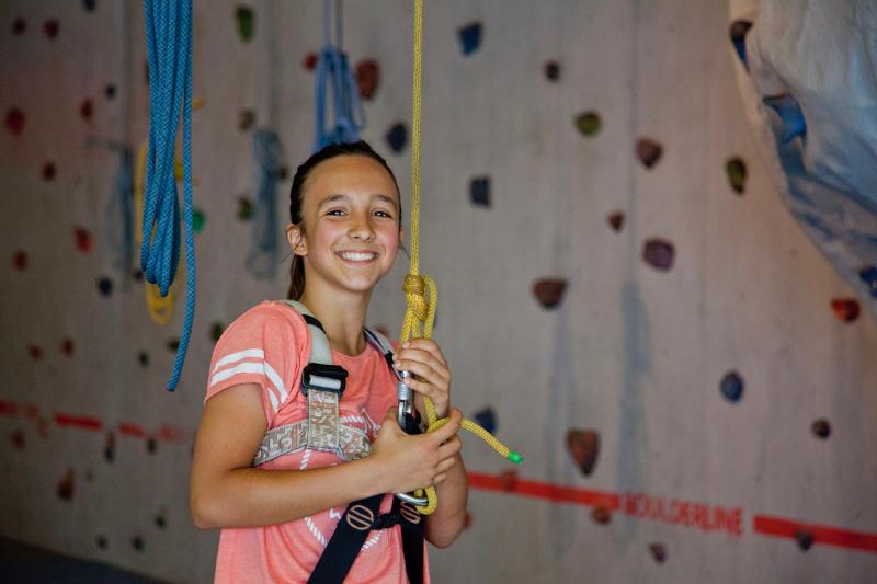 niña sonriendo en un rocódromo para hacer escalada con arnés y seguridad en un campamento de verano en aleman