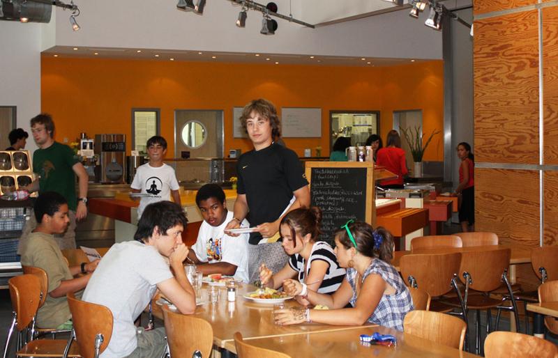 jovenes sentados en una mesa de un comedor o cafeteria comiendo con ropa de verano en un campamento aleman