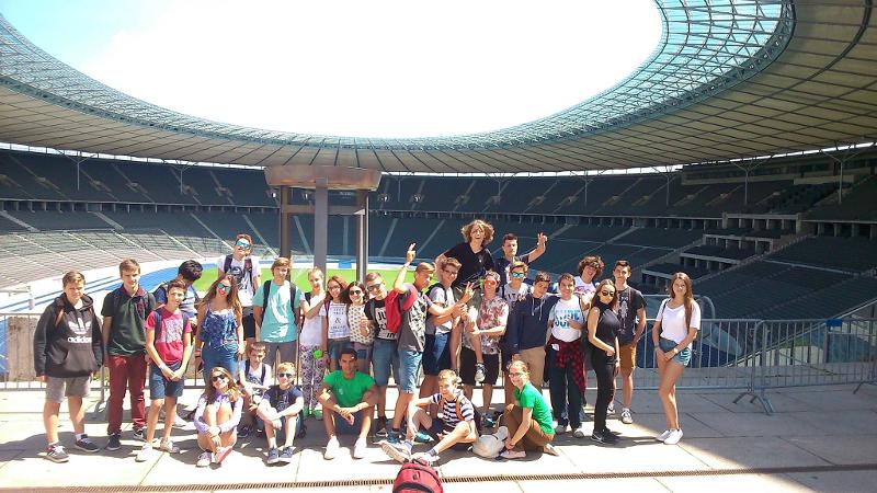 Grupo de niños y niñas visitando un estadio de fútbol posando bajo el sol en un campamento de verano en aleman