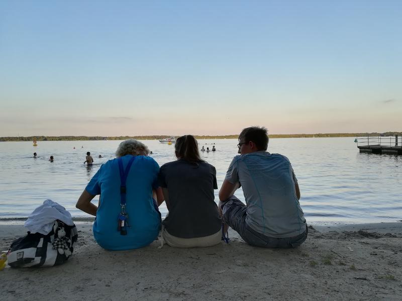 Tres amigos sentados juntos en la orilla de un lago viendo el atardecer mientras compañeros se bañan y juegan en el agua del campamento de verano en aleman