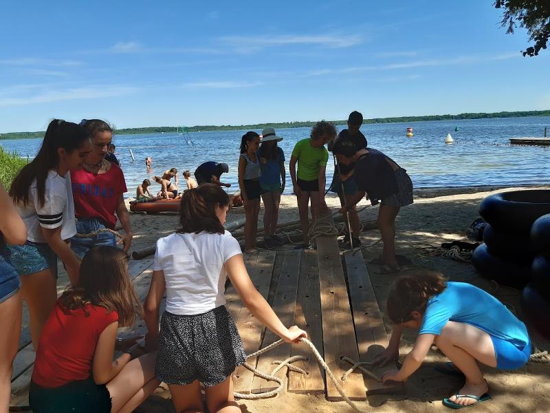 Jóvenes construyendo una balsa con madera y cuerdas en la orilla de un lago mientras compañeros se bañan en el agua en campamento en aleman
