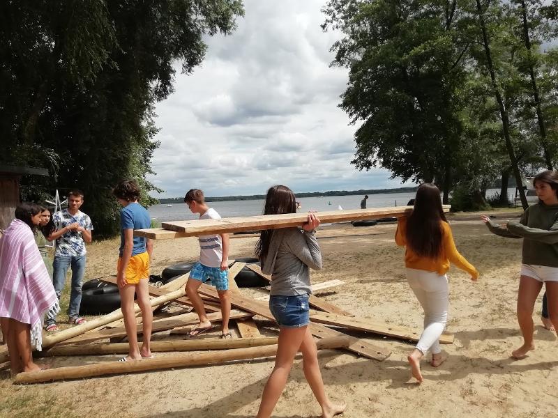 Jóvenes llevando tablas de madera para construir una balsa organizando los materiales en la orilla del lago en campamento alemania verano