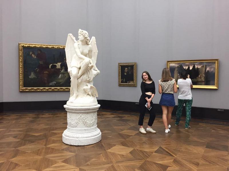 Chica posa en una sala de un museo de arte de la isla de los museos con cuadros y una escultura de un angel en curso verano alemania