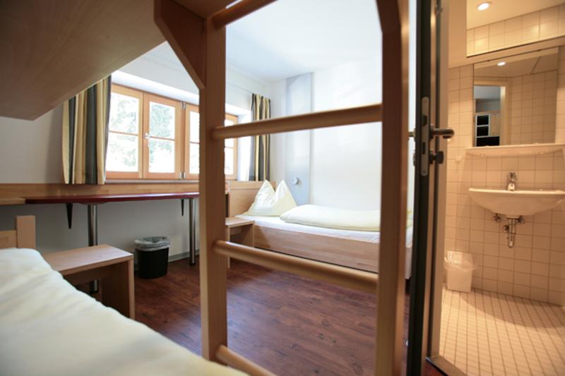 Habitación compartida con una litera y una cama individual con baño en suite y varias mesas frente a la ventana en curso de idiomas en alemania