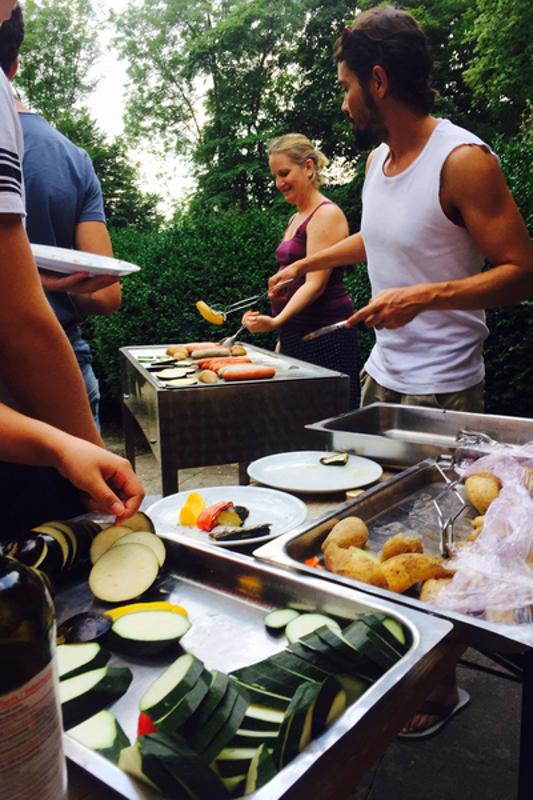 Monitores preparan salchichas y verduras en la barbacoa mientras campers esperan con platos en curso de idiomas en alemania
