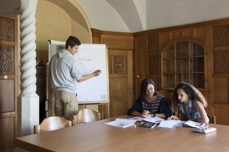 Dos amigas estudian sobre la mesa y un compañero explica cosas en la pizarra en una sala en curso de aleman en alemania