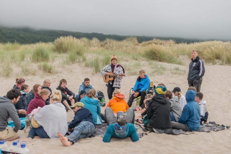 Jóvenes y sus monitores sentados en círculo mientras uno toca la guitarra en la playa