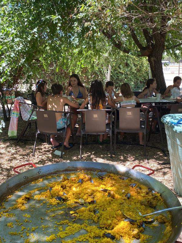 Niños y niñas comiendo paella en una mesa debajo de un árbol en un campamento para niños